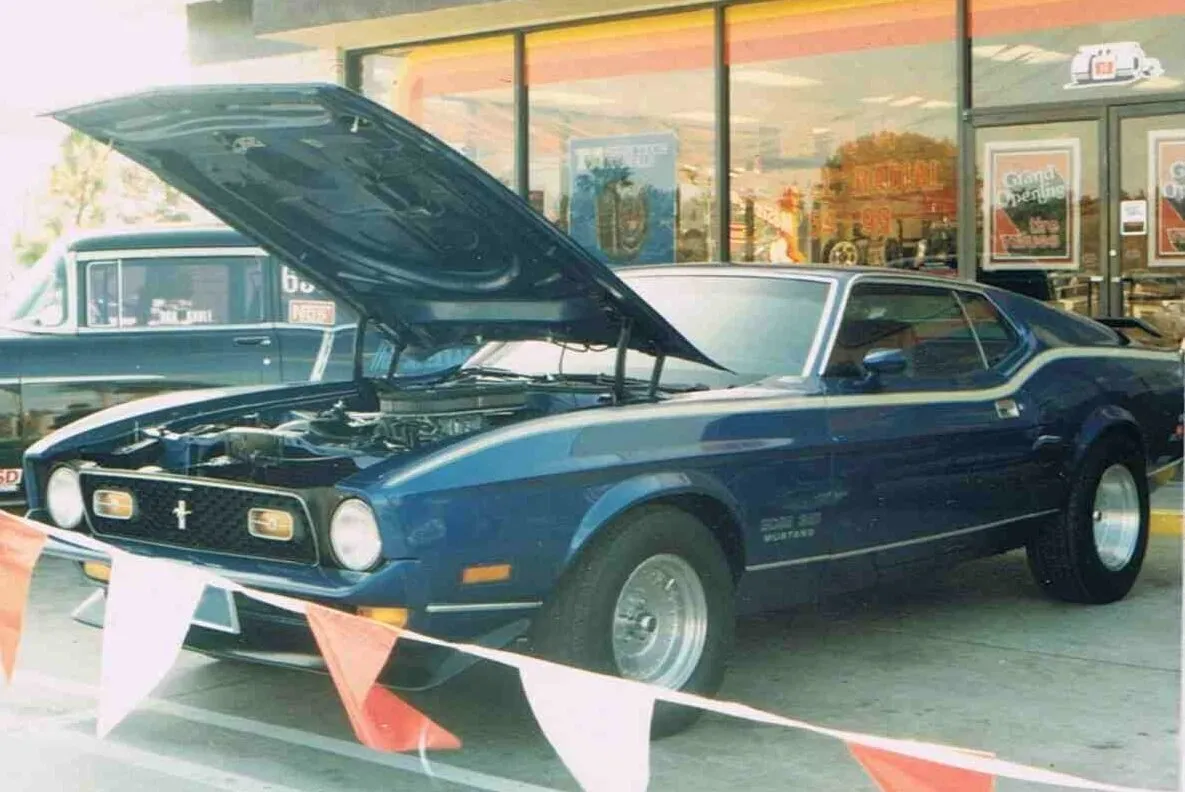 A dark blue Ford Mustang with its hood open, parked outside a store.